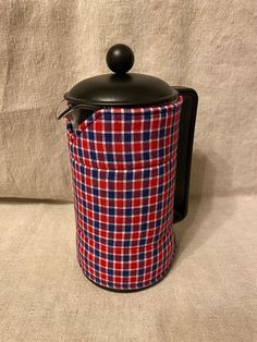 a red, white and blue plaid tea pot with a black lid sitting on a couch