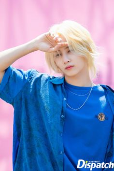 a man with blonde hair and blue shirt holding his hands up to his head while standing in front of a pink wall