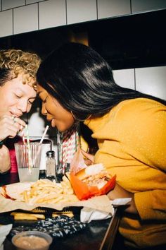 two people sitting at a table eating french fries and drinking sodas, with one woman looking on