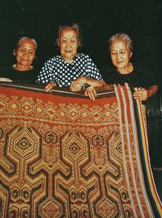 three older people are looking over an intricately designed blanket that is being held by two women