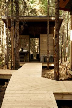 a wooden walkway leading to a small cabin in the woods with trees surrounding it and a table on one side