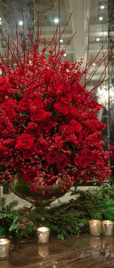 red flowers and greenery are arranged in a vase on the table next to candles