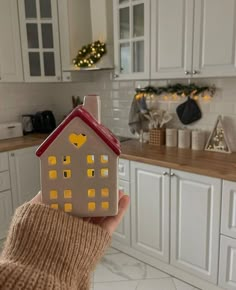 a person holding up a small house model in a kitchen with christmas lights on the cabinets