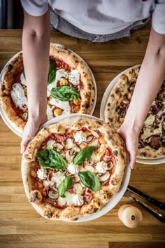 three pizzas with different toppings being held by two hands over a wooden table