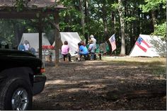 several people are gathered in the woods near tents with american flags on them and one person is sitting at a picnic table