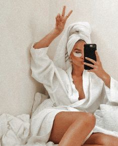 a woman in white robe sitting on bed holding up her cell phone to take a selfie