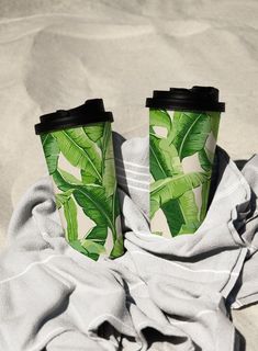 two coffee cups sitting on top of a blanket covered beach towel next to the ocean