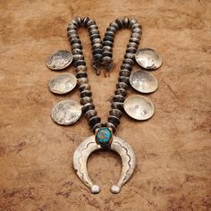 This Unique Heavy Old Pawn Squash Blossom Necklace Is A Stunning Example Of Navajo Jewelry Utilizing Mercury Dimes As Navajo Pearls And The Six Cupped Morgan Silver Dollar Coins Representing The Blossoms. The Morgan Dollars Date 1921 With The Top Left Dollar Date, 1892. The Horseshoe-Shaped Naja Pendant Features A Very Beautiful Oval-Shaped Dark Turquoise Gemstone, Set In A Bezel Style. The Pendant Hangs From A 26.5-Inch Silver Necklace, With What Appears As Hair Or Some Other Material, Along Wi Naja Necklace, Naja Pendant, Silver Dollar Coin, Turquoise Squash Blossom, Navajo Pearls, Morgan Dollars, Navajo Jewelry, Squash Blossom Necklace, Morgan Silver Dollar