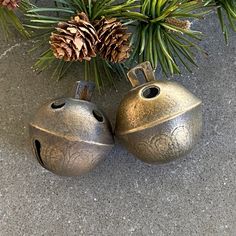 two metal bells sitting next to pine cones on top of a cement ground with evergreen needles