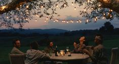 a group of people sitting around a table under a tree with lights strung over it