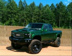 a green truck parked on top of a dirt road next to a field and forest