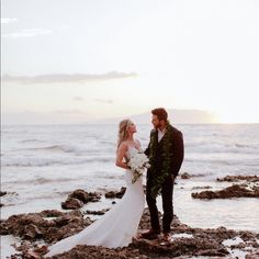 a bride and groom standing on the rocks by the ocean at sunset with their bouquet in hand