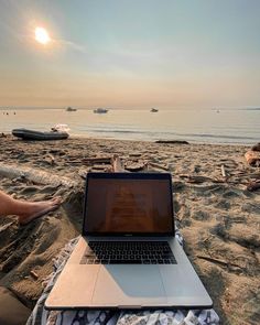 an open laptop computer sitting on top of a sandy beach next to the ocean with boats in the background