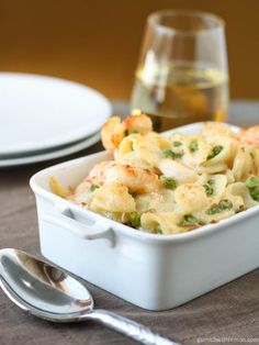 a white dish filled with pasta and shrimp next to a glass of wine on a table