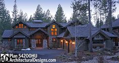 a large house with lots of windows and lights on it's front door, surrounded by pine trees