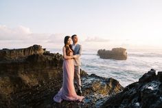 a man and woman standing on the rocks by the ocean with their arms around each other