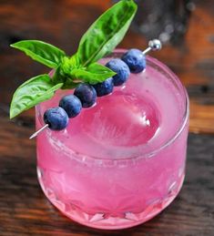 a pink drink with blueberries and green leaves on the rim, sitting on a wooden table