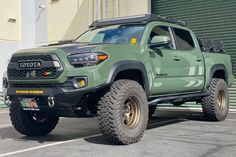 a green toyota truck parked in front of a building with large tires on it's flatbed