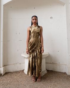 a woman standing in front of a white wall wearing a gold dress and sandals with her hands on her hips