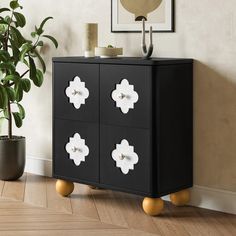 a black cabinet with four white flowers on the front and two gold legs, next to a potted plant