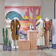 an assortment of women's clothing on display in front of a colorful painting and cactus