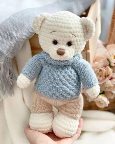 a crocheted teddy bear wearing a blue sweater and pants is held in someone's hand