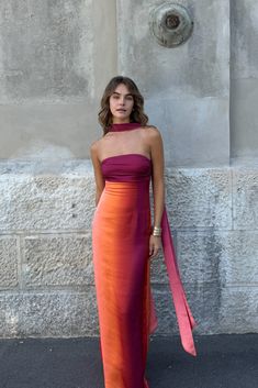 a woman standing in front of a wall wearing an orange and pink dress with a red sash around her neck