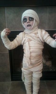 a little kid dressed up like an infant with bandages on his head and arms, standing in front of a fireplace