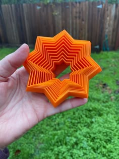 a hand holding an orange 3d object in the middle of a grassy area next to a fence