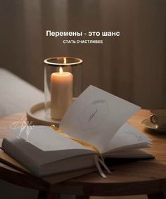 an open book sitting on top of a wooden table next to a candle and cup