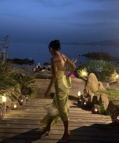 a woman in a yellow dress is walking down a wooden walkway by the water at night