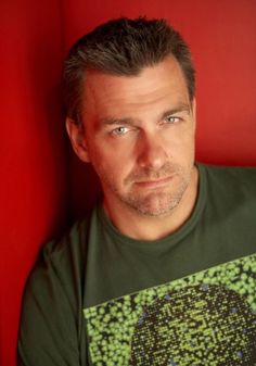 a man in a green shirt leaning against a red wall with his eyes wide open