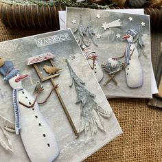 two cards with snowmen on them sitting next to a basket full of christmas decorations