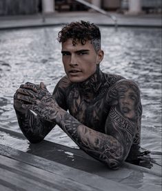 a man with tattoos sitting in the water next to a swimming pool and holding his hands together