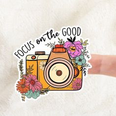 a hand holding up a sticker that says focus on the good with an image of a camera and flowers