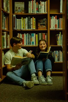 a man and woman sitting on the floor in front of a bookshelf reading