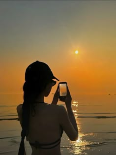 a woman standing on the beach taking a photo with her cell phone at sunset or dawn