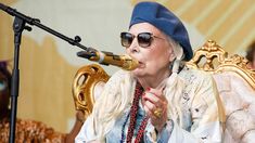 an older woman wearing sunglasses and a blue hat is sitting in front of a microphone