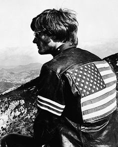 a man sitting on top of a mountain with an american flag jacket over his shoulders