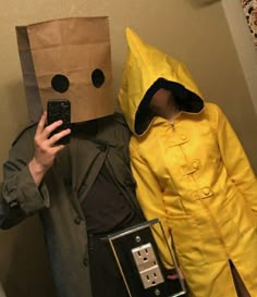 two people dressed in costumes standing next to each other, one wearing a paper bag and the other holding a cell phone