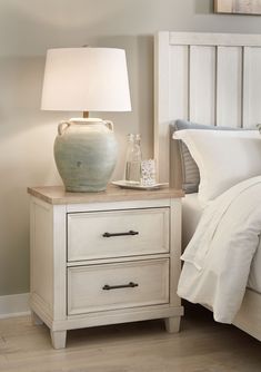 a nightstand with two drawers and a lamp on it next to a bed in a bedroom