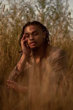 a man with dreadlocks sitting in tall grass talking on a cell phone while holding his hand to his ear