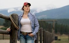 a woman standing next to a wooden fence with mountains in the backgrouds