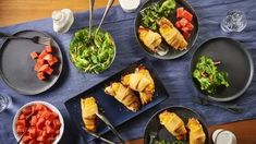 several plates with food on them sitting on a blue table cloth next to glasses and utensils