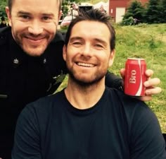 two men are smiling and posing for the camera with one holding a can of soda