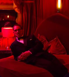 a man in a tuxedo sitting on a bed with red lights behind him