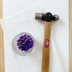 purple flowers in a glass bowl next to a hammer and paper towel on a wooden table