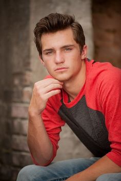 a young man poses for a photo in front of a brick wall with his hand on his chin