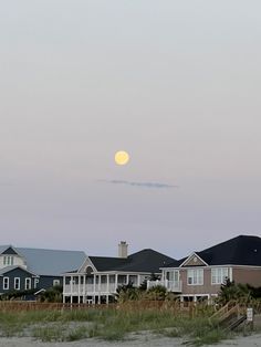 a full moon is seen over houses on the beach in front of some sand dunes