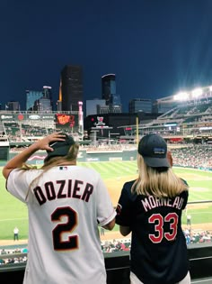 two women in baseball uniforms are looking at the stadium from behind home plate with lights on